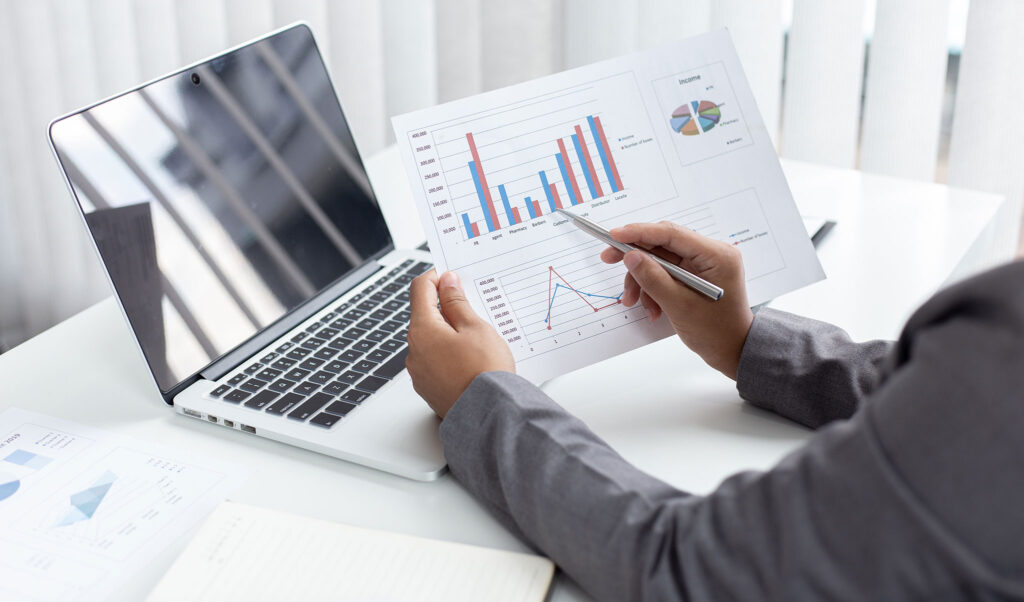 homem com folha na mao mostrando grafico sobre finanças com notebook aberto na mesa