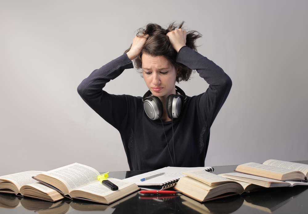 mulher com fone de ouvido no pescoço e segurando o cabelo com raiva e com diversos livros a sua frente