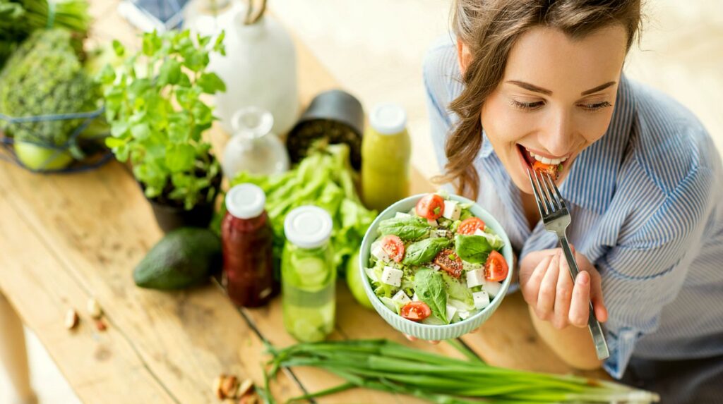 mulher comendo salada ao lado de uma mesa com diversos legumes
